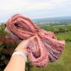 A hand wearing a white watch holds up a thick skein of multicolored, handspun yarn in shades of pink, orange, and purple and blue. The handspun yarn is called "Adventures of Indie Dyeing 2.1." The background shows a green grassy landscape, distant trees, and a cloudy sky, with “LEILANI HANDMADE” faintly visible at the bottom.