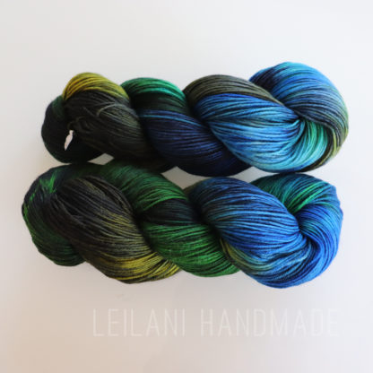 Two skeins of yarn are displayed against a white background. The yarn features a gradient of colors including blue, green, yellow, and black. "LEILANI HANDMADE" is lightly visible in the background at the bottom.