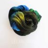 A skein of yarn is coiled in a loose spiral, displaying vibrant shades of black, green, yellow, and blue. The background is white, and the faint text “LEILANI HANDMADE” is visible near the bottom of the image.