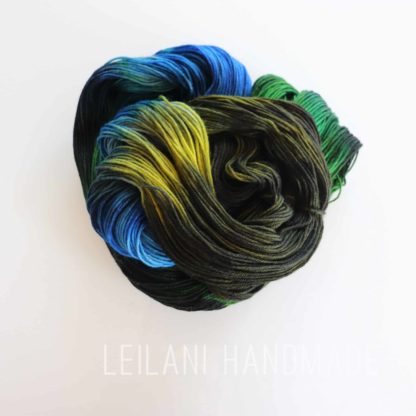 A skein of yarn is coiled in a loose spiral, displaying vibrant shades of black, green, yellow, and blue. The background is white, and the faint text “LEILANI HANDMADE” is visible near the bottom of the image.