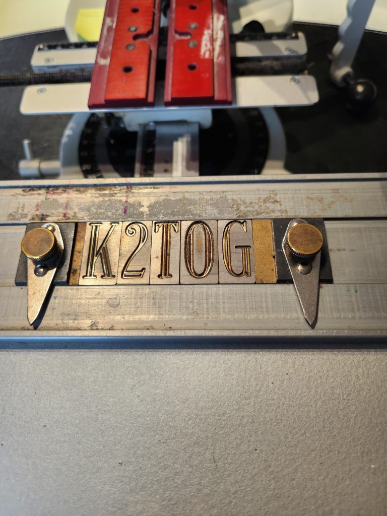 brass fonts spelling out K 2 T O G on a metal tray of a pantograph engraver