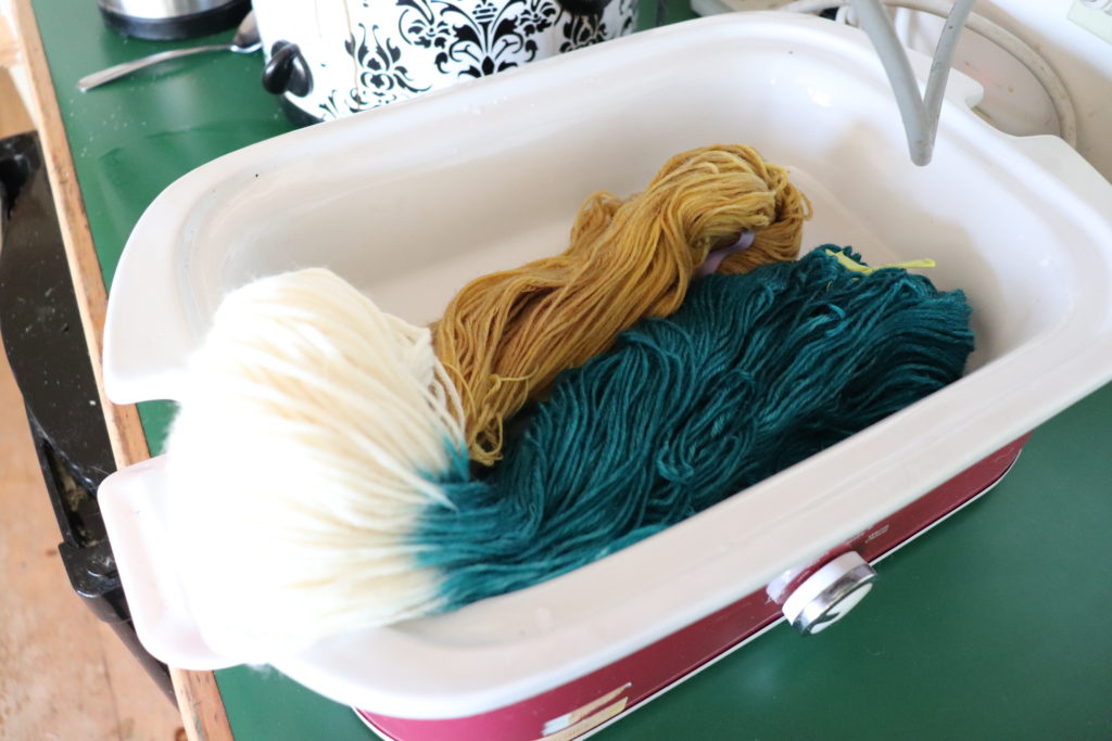 an open rectagular crockpot displaying 2 skeins of yarn tied together with two ends dyed. One end golden, the other end teal