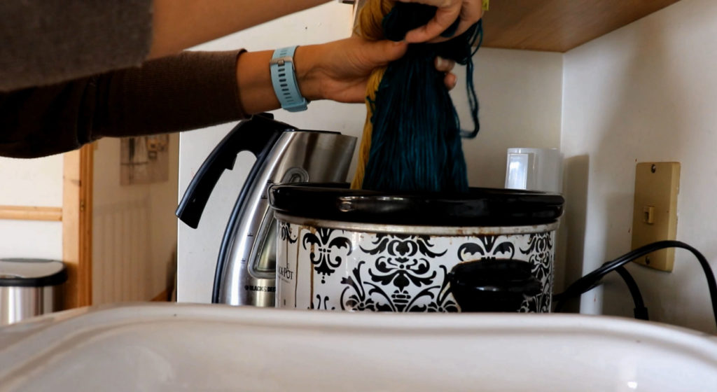 hands dipping a section of yarn into a black and white crockpot while holding up the teal and golden sections