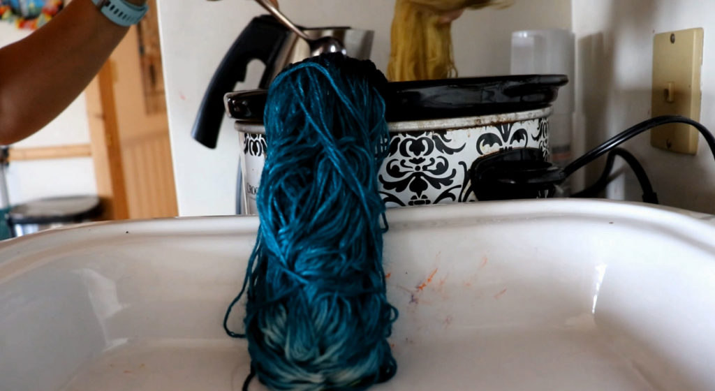a section of teal colored yarn hanging outside a crockpot while a hand works a spoon into the liquid in the corckpot, the other hand holding up the golden section of yarn