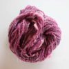A skein of handspun yarn in varying shades of pink, purple, and hints of white is coiled into a loose bundle. The yarn has a thick and thin texture, giving it a slightly uneven, handcrafted appearance. The background is plain and light.