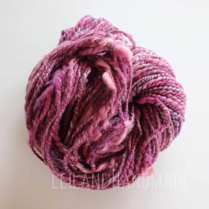 A skein of handspun yarn in varying shades of pink, purple, and hints of white is coiled into a loose bundle. The yarn has a thick and thin texture, giving it a slightly uneven, handcrafted appearance. The background is plain and light.
