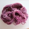 A skein of handspun yarn in varying shades of pink, purple, and hints of white is coiled on a white surface. The yarn has a soft, slightly fuzzy texture and a twisted, plied appearance. The words “LEILANI HANDMADE” are faintly visible underneath.