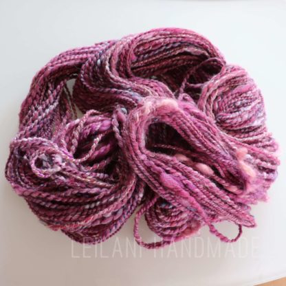 A skein of handspun yarn in varying shades of pink, purple, and hints of white is coiled on a white surface. The yarn has a soft, slightly fuzzy texture and a twisted, plied appearance. The words “LEILANI HANDMADE” are faintly visible underneath.