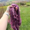 A hand wearing a white watch holds up a bundle of chunky, pink and purple handspun yarn outdoors, with a green lawn, scattered dandelions, a wooden fence, and a scenic countryside landscape in the background.