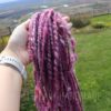 A hand holding a bundle of thick, handspun, pink and purple yarn outdoors, with a grassy yard and distant landscape in the background. The person is wearing a light-colored watch. Text at the bottom reads “LEILANI HANDMADE.”.