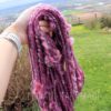 A hand holds several strands of thick, textured pink and purple yarn outdoors. The background shows a grassy yard with dandelions, plants, a fence, and a distant hilly landscape under a cloudy sky. The yarn appears soft and curly.