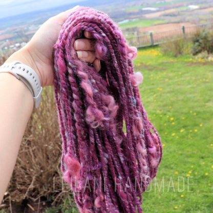 A hand holds several strands of thick, textured pink and purple yarn outdoors. The background shows a grassy yard with dandelions, plants, a fence, and a distant hilly landscape under a cloudy sky. The yarn appears soft and curly.