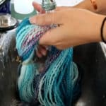 A person holds blue and purple yarn under running water from a kitchen faucet, rinsing it in a stainless steel sink. The hands, one with a black hair tie and the other with a copper bracelet, grip the yarn as water flows over it.