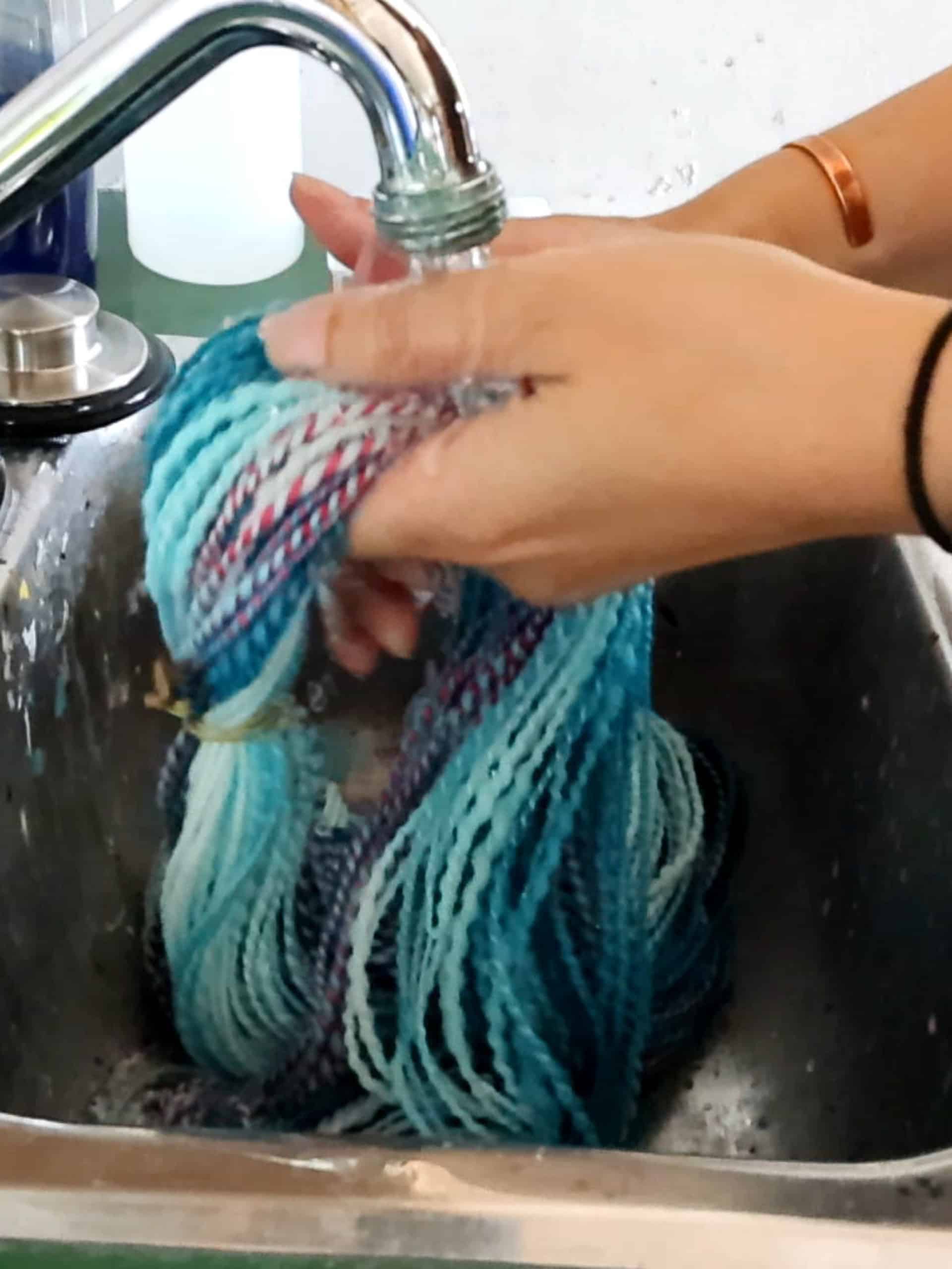 A person holds blue and purple yarn under running water from a kitchen faucet, rinsing it in a stainless steel sink. The hands, one with a black hair tie and the other with a copper bracelet, grip the yarn as water flows over it.