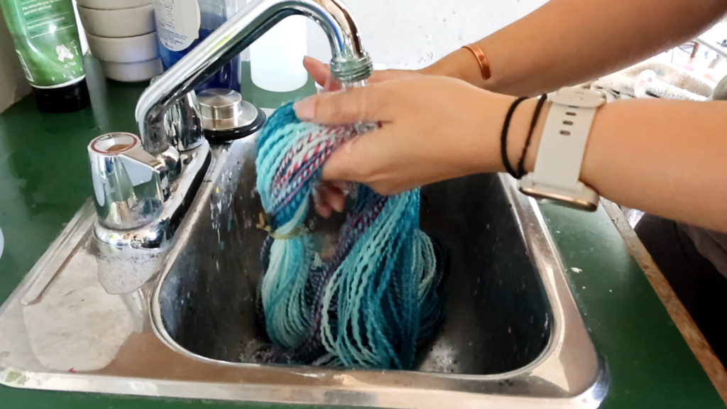 A person wetting a skein of blue and multicolored yarn  under a running tap in a kitchen sink for setting the twist. Their hands are holding the yarn, and their wrists have a watch and bracelets. Various containers and items are visible around the sink.