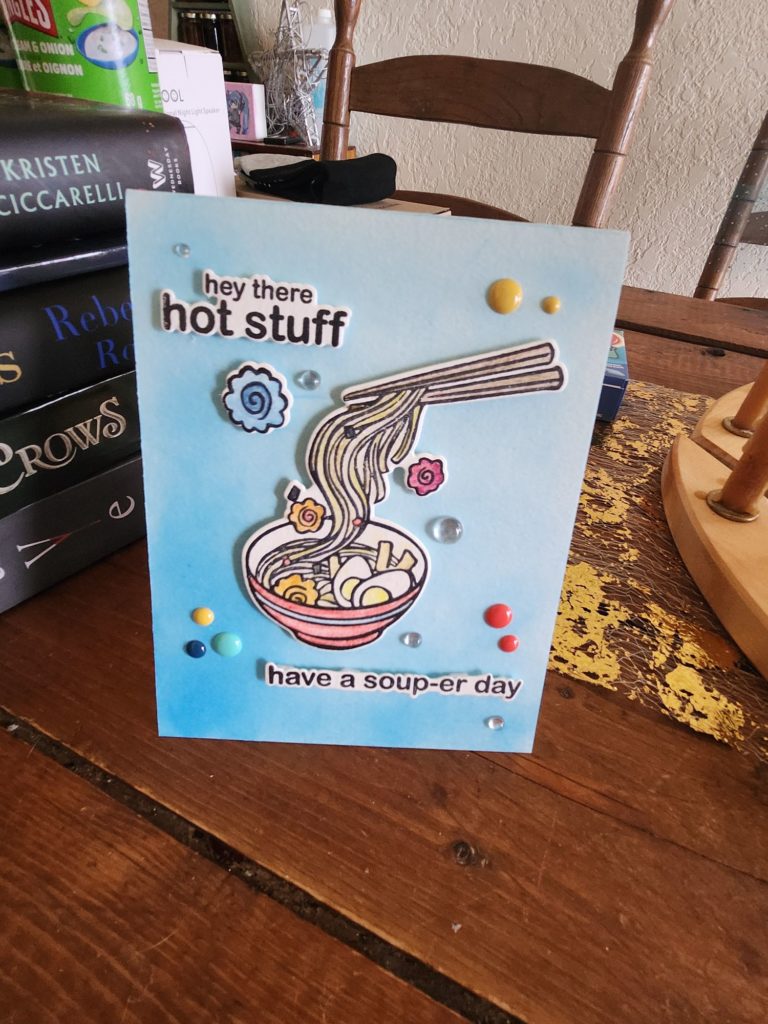 A colorful greeting card is displayed on a wooden table. The card features an illustration of a bowl of noodles with chopsticks and decorative elements. Text on the card reads "hey there hot stuff" at the top and "have a soup-er day" at the bottom. Books and other items are visible in the background.