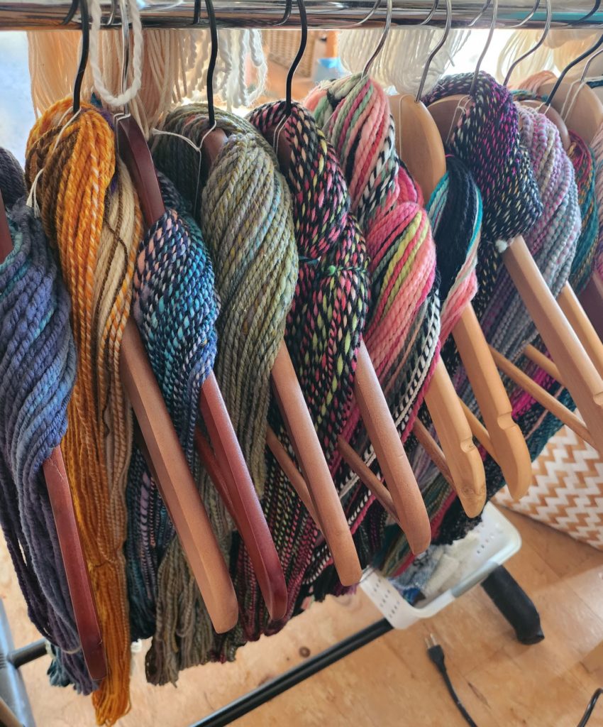 Several skeins of colorful yarn are hanging on wooden hangers. Each skein displays a mix of vibrant hues, including shades of blue, green, yellow, pink, and purple. The yarns are neatly organized and ready for crafting. The background shows a wooden floor.