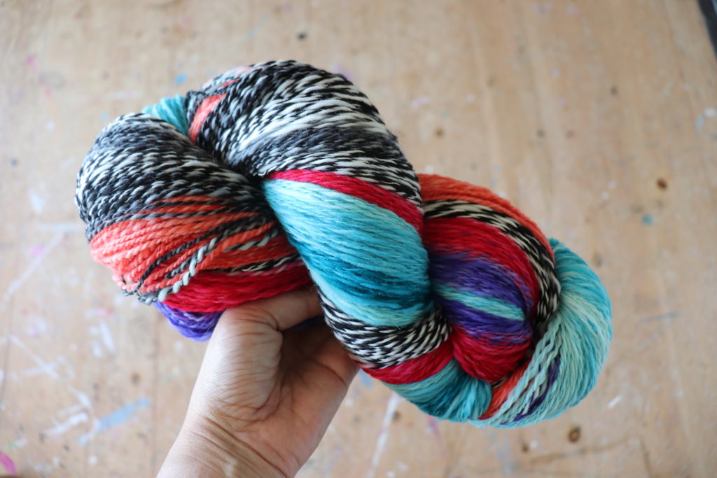 A hand holding a twisted skein of colorful yarn with shades of red, teal, black, white, and purple. The background is a wooden surface.