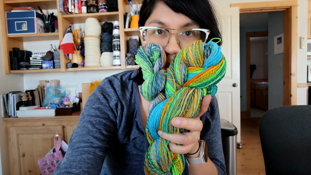 A person with glasses holds colorful skeins of yarn in front of their face. They are in a room with shelves filled with books and more yarn in the background. The yarn is vibrant with green, blue, yellow, and orange hues. Join me as I spin this yarn in the above video.