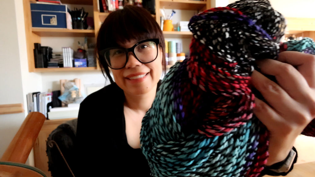 A person with glasses and short hair smiles while holding colorful, patterned crepe yarn. Books and various items are visible on shelves in the background.