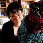 A person with glasses and short hair smiles while holding colorful, patterned crepe yarn. Books and various items are visible on shelves in the background.