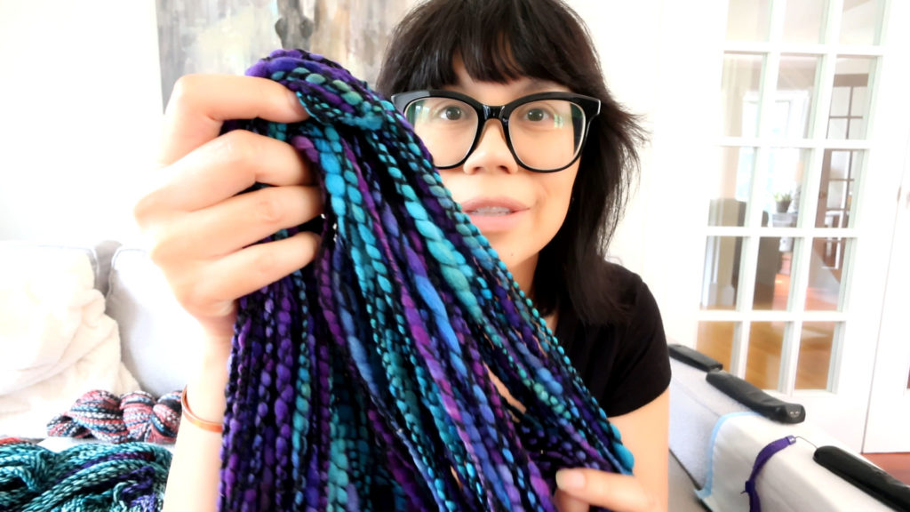 A person with glasses holds up strands of braided yarn in vibrant shades of blue, purple, and teal. They are indoors, wearing a black shirt, with a couch and bright room in the background.