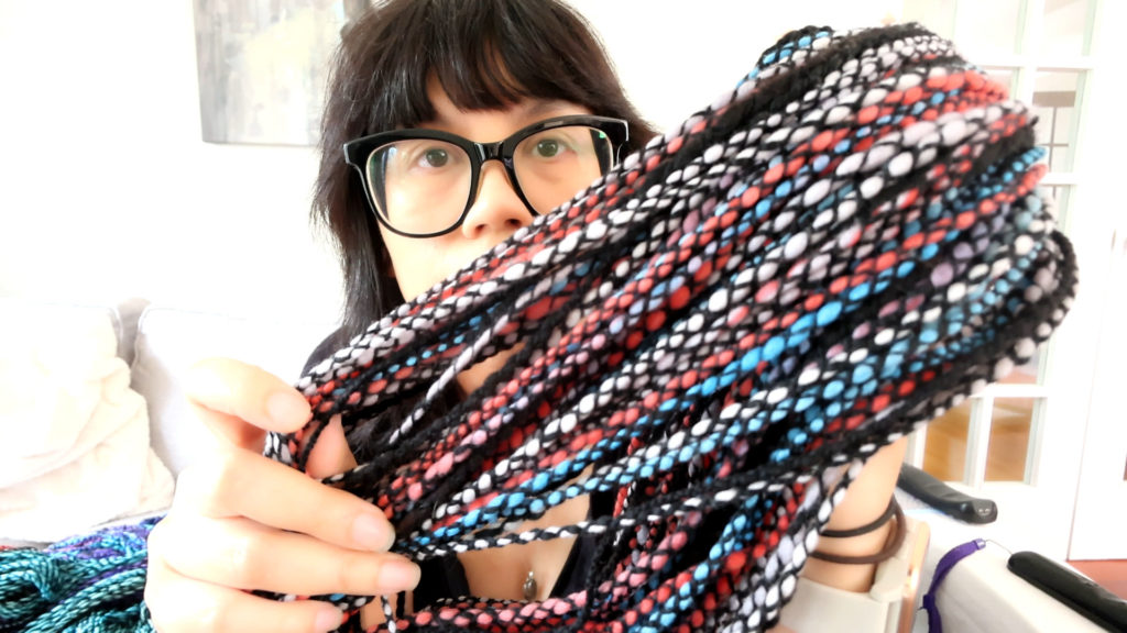 A person with glasses holds colorful braided yarn close to the camera, partially obscuring their face. The yarn consists of vibrant strands in red, blue, black, and white. The background features a light interior setting.