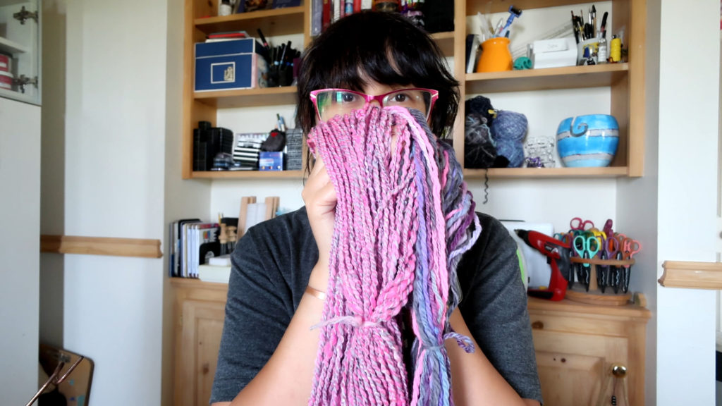 Person holding pink and purple yarn in front of their face, standing in a room with shelves filled with books, craft supplies, and decorations in the background.