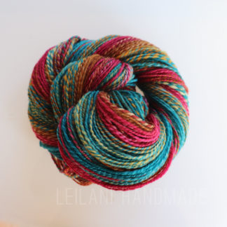 A skein of multicolored yarn featuring vibrant strands of teal, red, and gold twisted together. The fibers create a textured and visually striking pattern. The words "Leilani Handmade" are faintly visible in the background.