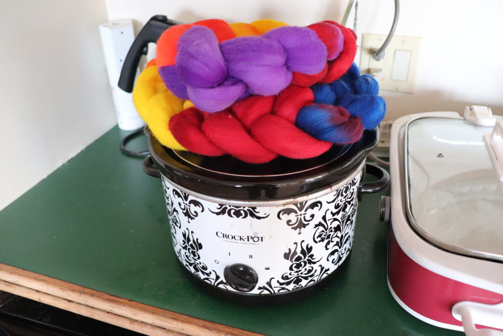 A crockpot with a decorative black and white pattern has a braid of colorful wool roving in shades of red, orange, yellow, blue, and purple, sits on top, placed on a green countertop next to another red and white crockpot.