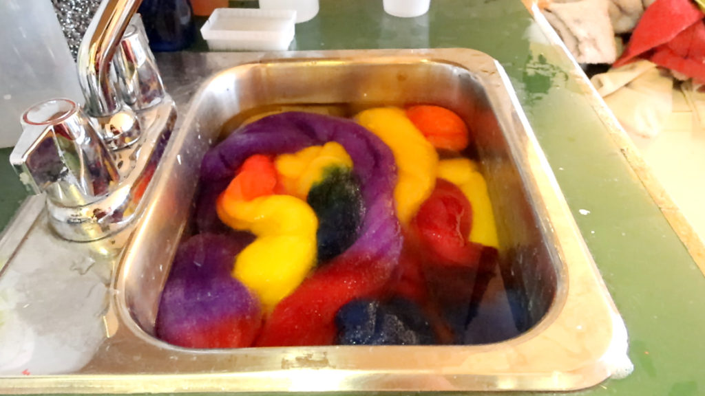 Colorful wool roving soaking in a metal sink filled with water. Various shades of purple, yellow, orange, red, and green are visible, after a fiber dyeing process. A silver faucet is on the left side of the sink.