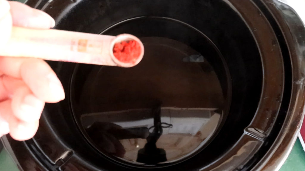 A hand holds a white one quarter teaspoon containing red powder used to dye roving in a black slow cooker filled with water. The background is slightly blurred, focusing on the spoon and the cooker.