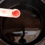 A hand holds a white one quarter teaspoon containing red powder used to dye roving in a black slow cooker filled with water. The background is slightly blurred, focusing on the spoon and the cooker.