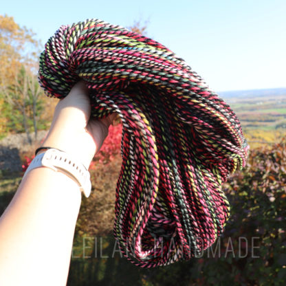 A person holds a skein of multicolored yarn against a scenic outdoor backdrop of trees and a distant landscape. The yarn features shades of pink, black, green, and white.