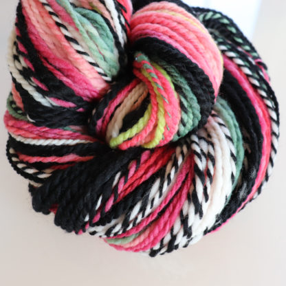 A skein of handspun merino yarn featuring a vibrant mix of colors including pink, black, white, green, and yellow, twisted together in a spiral pattern. The texture is soft and the fibers appear plush.