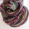 A close-up of a skein of colorful, twisted handspun merino yarn featuring shades of pink, green, and white, accented by a strand of black, artistically coiled on a white surface.