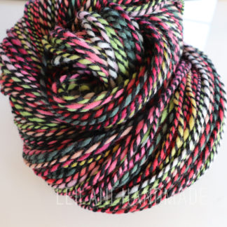 A close-up of a skein of colorful, twisted handspun merino yarn featuring shades of pink, green, and white, accented by a strand of black, artistically coiled on a white surface.