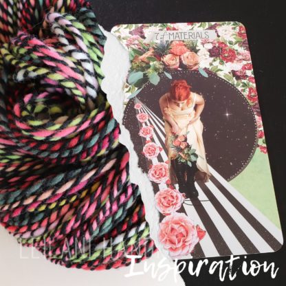 A colorful, handspun yarn beside a card depicting a woman on a striped pathway surrounded by roses and a cosmic background. The card is titled "7 of Materials." The word "Inspiration" is written at the bottom.