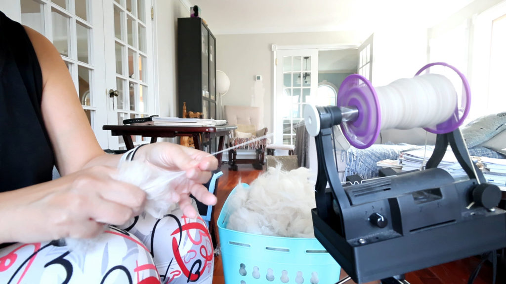 A person spins yarn on an e-spinner spinning machine in a bright, cozy room. They hold a bundle of white fiber, with more fiber in a blue basket nearby. Natural light fills the space, highlighting a table and furniture in the background.