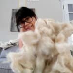 A person with glasses and dark hair sits on a couch, smiling while holding up a large pile of fluffy, white wool. The background includes a gray couch, a lamp, and a door with glass panes.