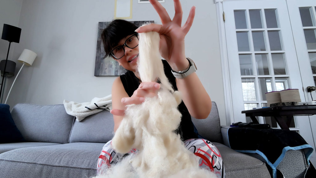 A person with glasses sits on a sofa, holding a piece of fluffy fiber. They're engaged in a fiber craft, with their hands in focus. The room has a cozy vibe, with a lamp and framed pictures visible in the background.