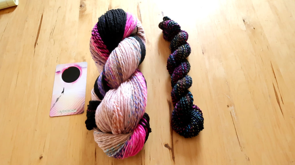 Two yarn skeins on a wooden table: left skein is thick, multicolored with pink, cream, black; right skein is smaller, tightly twisted with black and colorful speckles. A card with a pink and black design lies beside them. The card was used for designing yarn. 