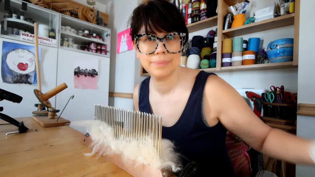 A person wearing large glasses is seated at a wooden table working with wool with a hackle blending tool that has a row of metal prongs. Shelves behind them hold various yarns and craft materials. The person is smiling slightly and wearing a dark sleeveless top.