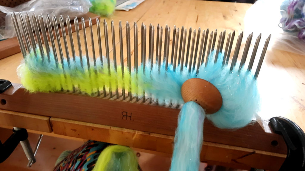 A wooden blending hackle with sharp metal tines is clamped to a table. Vibrant strands of yellow, green, and blue wool fibers are blended together, while a strand of pale blue wool threads through a round wooden diz. A wooden blending hackle with sharp metal tines is clamped to a table. Vibrant strands of yellow, green, and blue wool fibers are blended together, while a strand of pale blue wool threads through a round wooden diz.