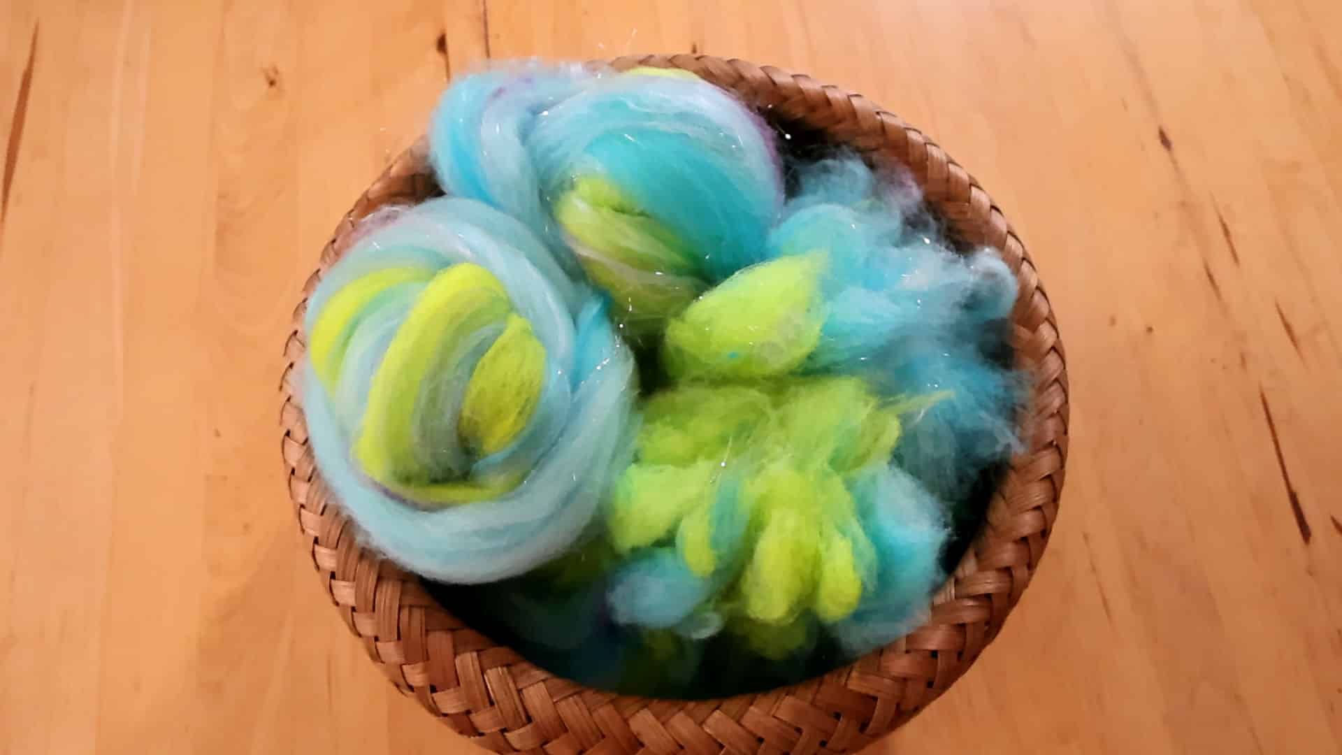 A woven basket filled with fluffy wool roving in shades of blue, green, and yellow, sitting on a wooden surface.