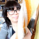 A person wearing glasses and a gray sweater smiles while holding up strands of yarn in various colors, including yellow, orange, and cream. Shelves with books and boxes are visible in the background.
