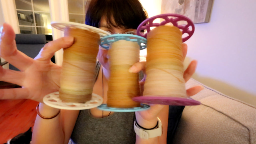 A person holds up three large spools of yarn in shades of brown, each with a different colored base: white, blue, and purple. The background shows a cozy living room with a couch and windows.
