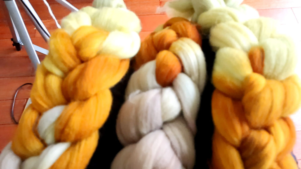 Close-up of three thick braids made from wool roving in deep colors, including shades of yellow, orange, and cream. They are arranged side by side on a wooden surface, creating a textured and colorful display. These rovings are for creating yarn.