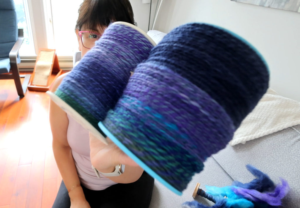A person holds up two large spools of handspun yarn in shades of blue and purple, with their face partially obscured. Yarn fibers and a spinning wheel are visible in the background.