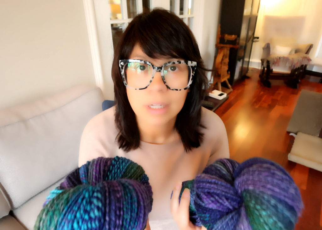 A person with dark hair and large glasses holds two skeins of multicolored superwash BFL yarn, featuring blue, purple, and green shades, while sitting on a couch in a cozy, well-lit living room.